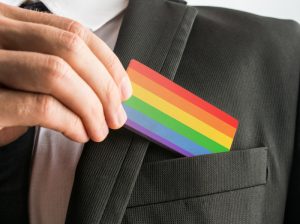 Man withdrawing a wooden card painted as the gay pride flag from his suit pocket, close up of his hand.